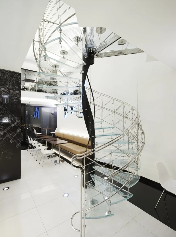 The glass and steel spiral staircase standing in the Café B. Braun interior, with the dark tapering steel newel at its centre and a lattice of thin stainless rods around it